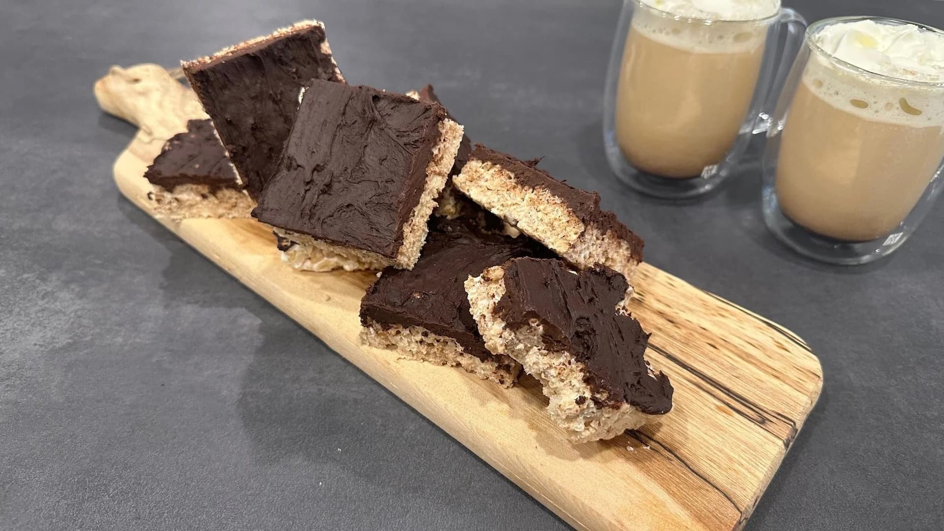 Chocolate-topped protein rice krispies treats cut into squares on a wooden board with coffee drinks beside them.