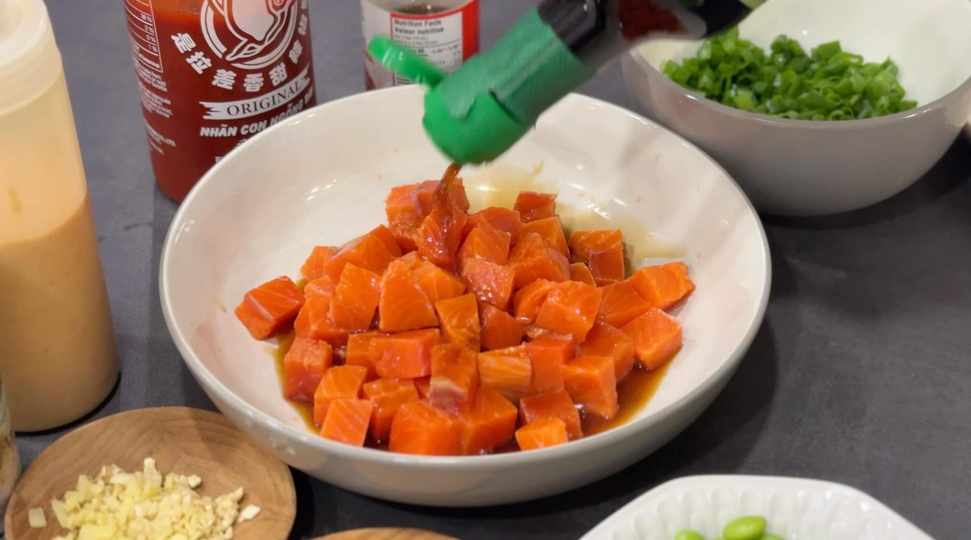 Diced salmon mixed with soy sauce and seasonings in a white bowl, surrounded by prep ingredients including green onions and sriracha.