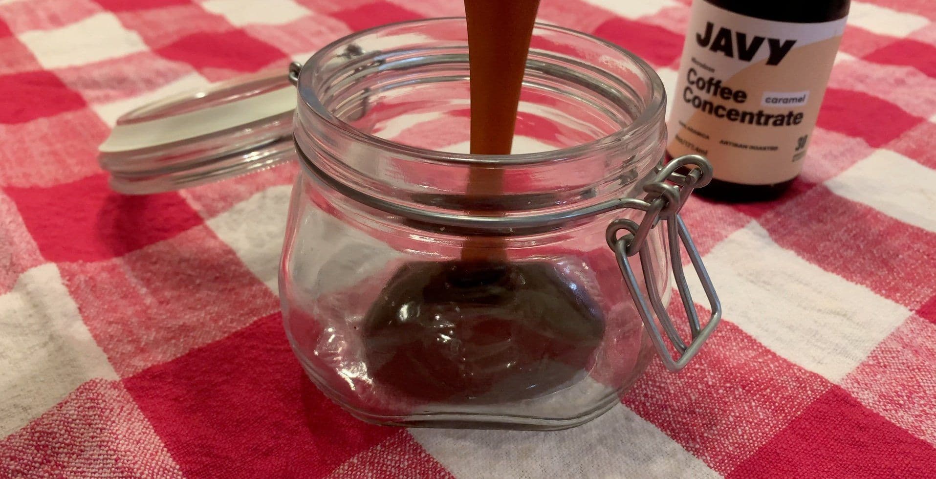 Whiskey caramel sauce being made in a glass jar with a wooden spoon, with JAVY coffee concentrate bottle nearby on a red gingham cloth.
