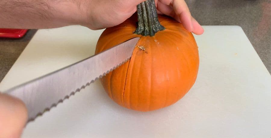 Cutting a small pumpkin in half with a serrated knife on parchment paper.
