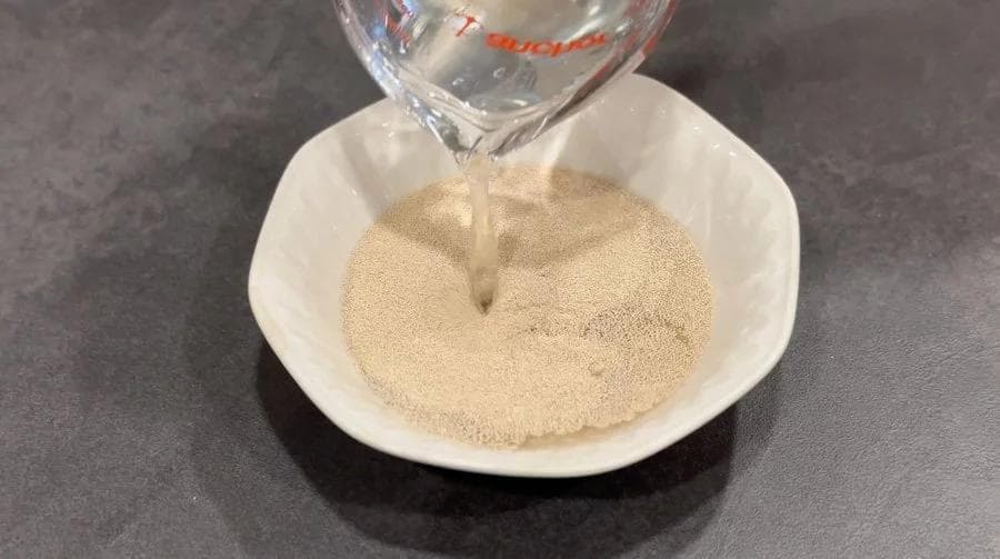 Lukewarm water being poured into a bowl of yeast and sugar mixture for pita bread dough.