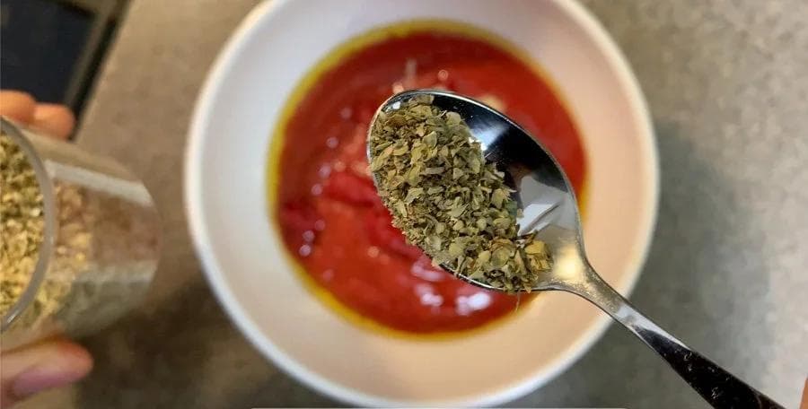 Spoon of dried oregano being added to a bowl of red pizza sauce with olive oil drizzle.