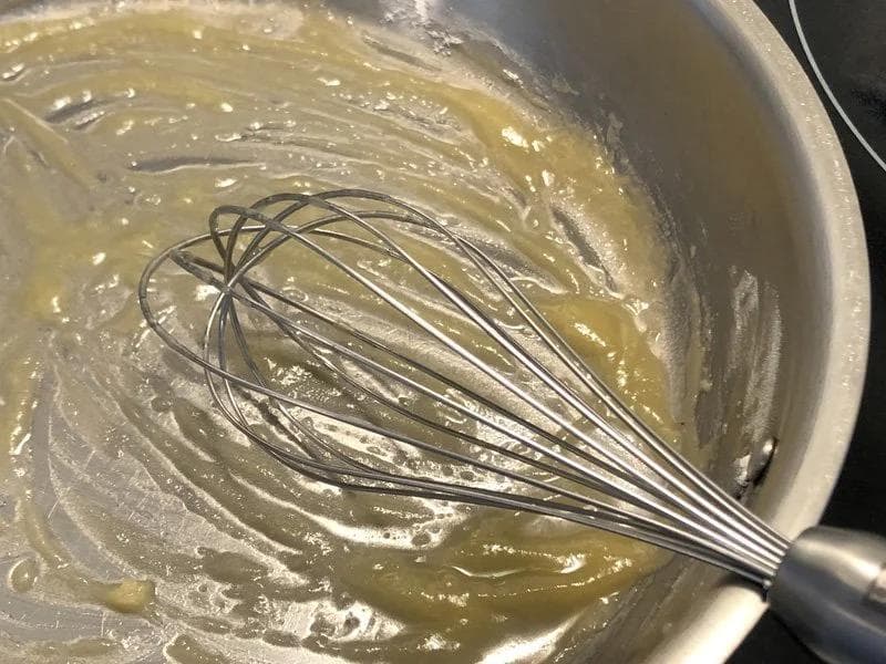 Whisking butter and flour roux in a white pot, creating a smooth pale mixture for béchamel sauce.