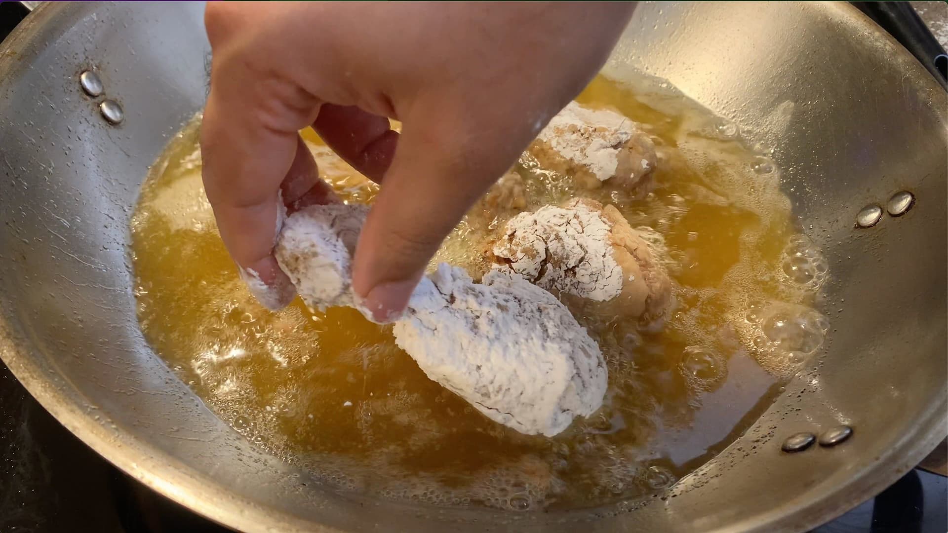 Breaded chicken drumsticks being placed into hot oil for deep frying in a stainless steel pan.