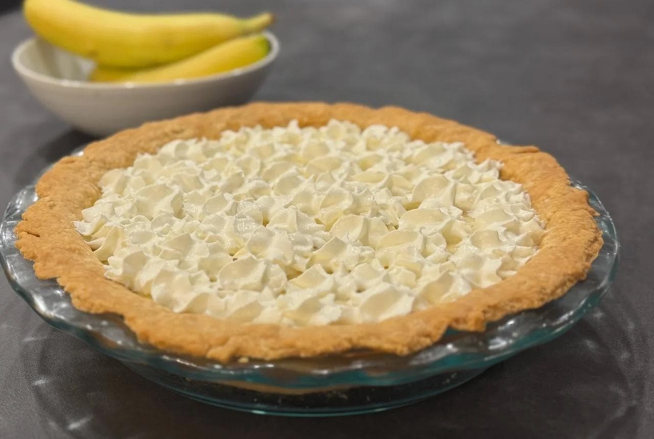 Banana cream pie with graham cracker crust and whipped cream topping in a glass pie dish, with bananas in a bowl behind it.