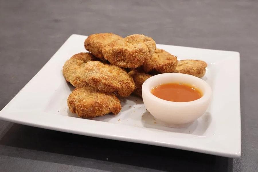 Golden air fryer chicken nuggets served on a white plate with a small bowl of dipping sauce.