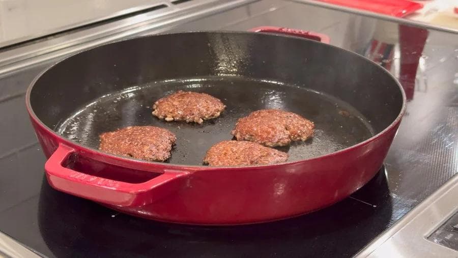 Three browned ground turkey patties sizzling in a red skillet on a stovetop during cooking.