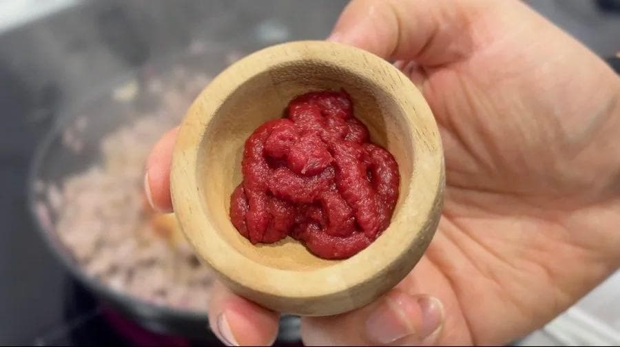 Tomato paste in a wooden measuring bowl held in a hand over a pot of ground beef.