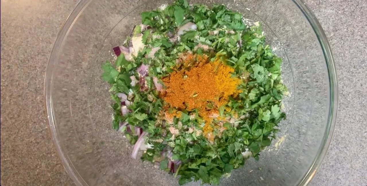 Dry ingredients for besan chilla mixed in a glass bowl: chopped cilantro, red onion, turmeric powder, and spices.