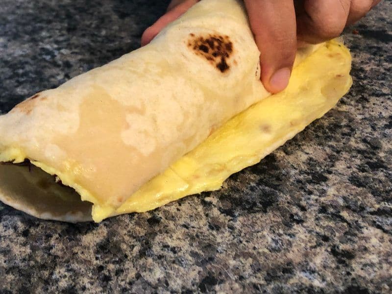Hands wrapping a chicken kathi roll with yellow paratha and filling on a granite countertop.