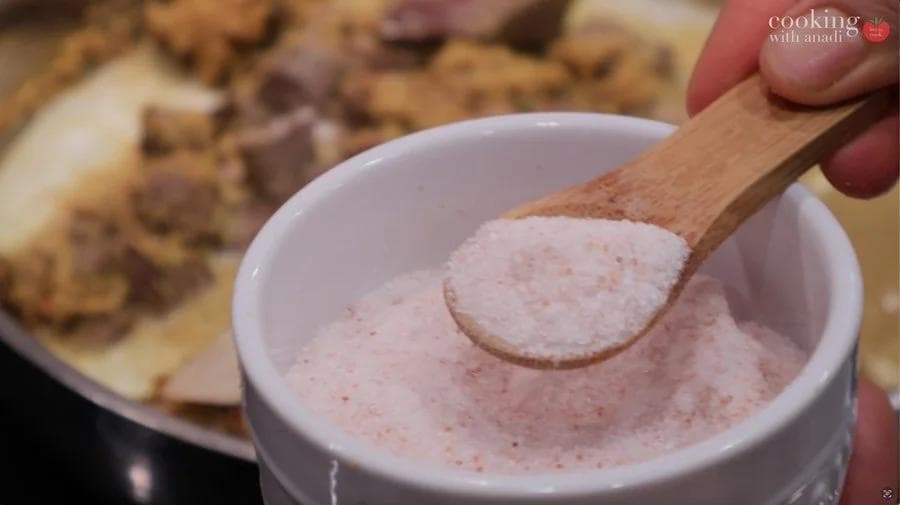 Wooden spoon holding a scoop of pink-hued spice mixture over a white bowl during Balinese beef rendang preparation.