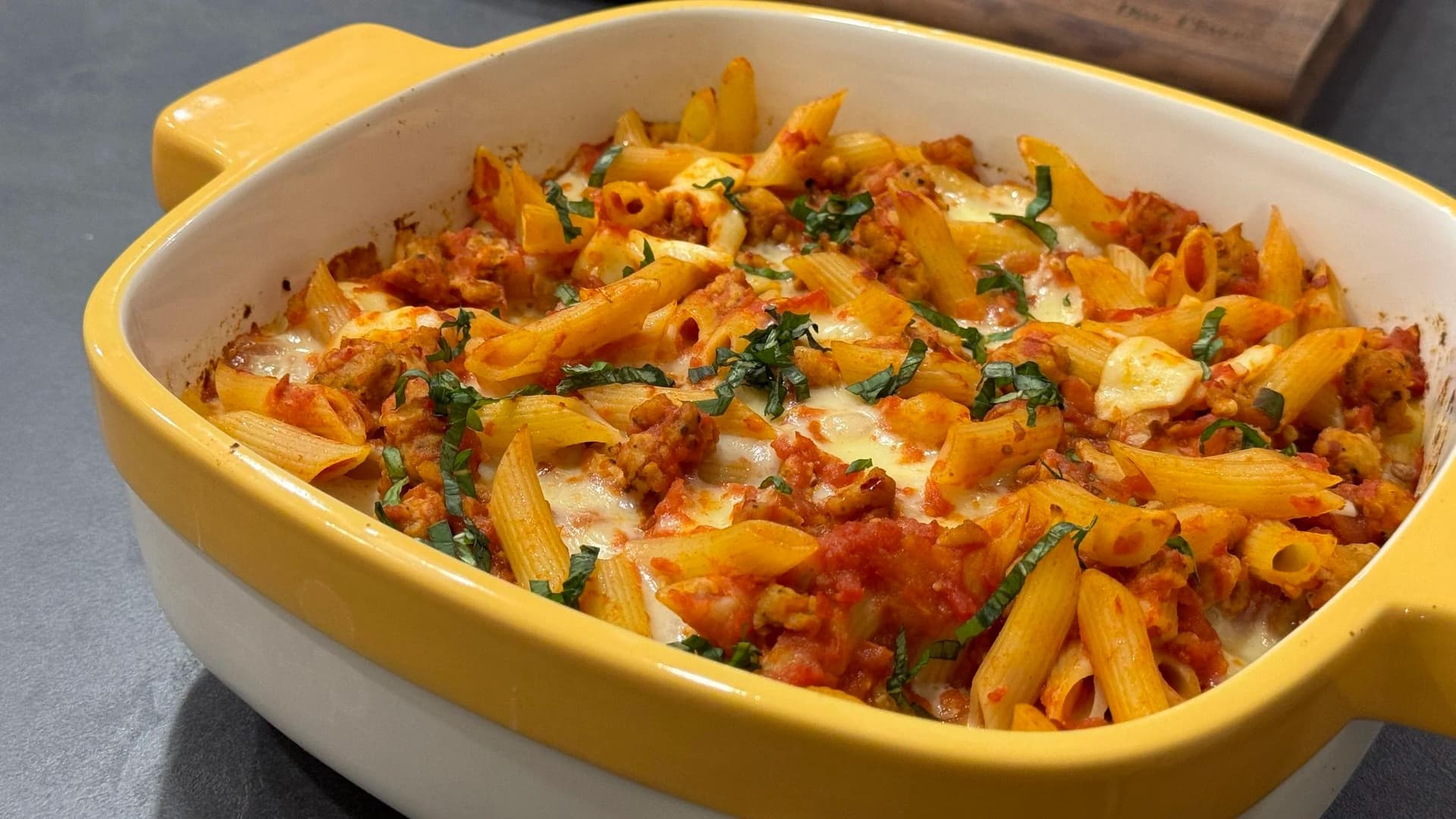 Cheesy baked penne with spicy Italian sausage in a yellow baking dish, topped with melted cheese and fresh basil.