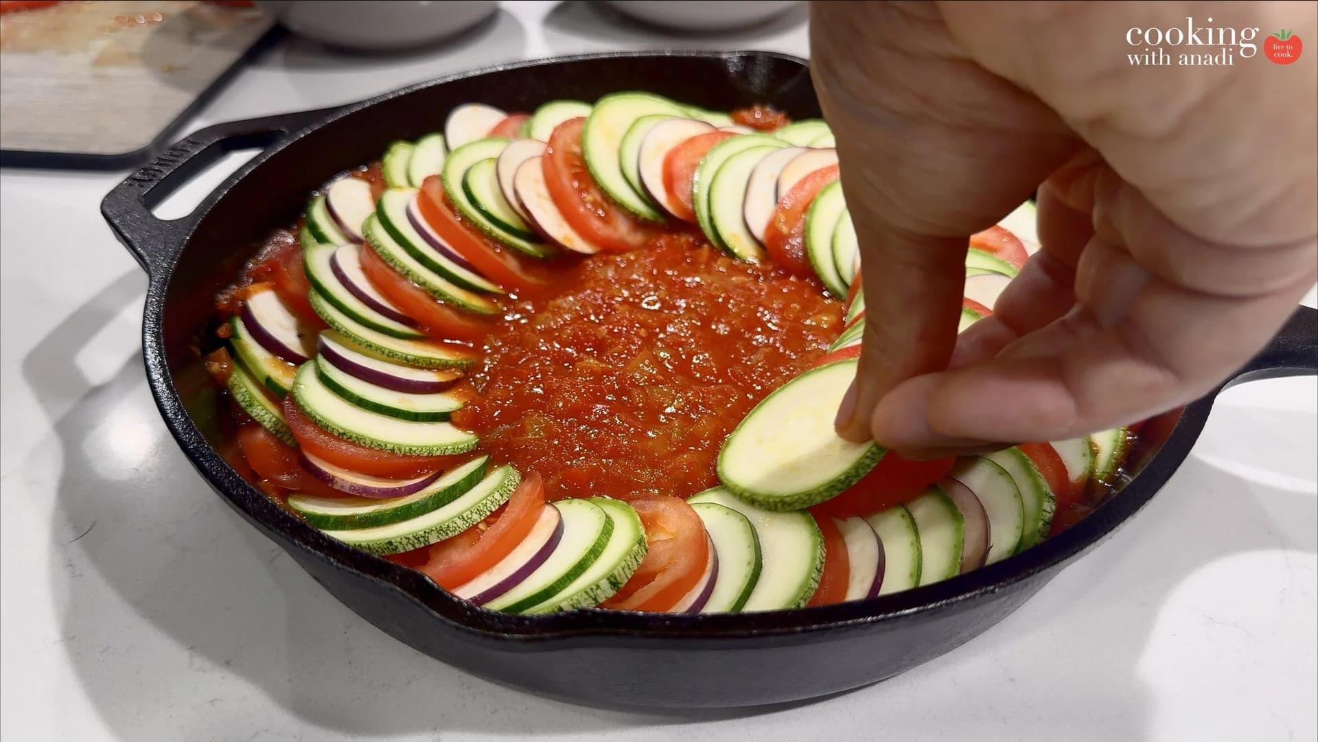 Sliced zucchini, tomato, and red onion arranged in overlapping spirals over tomato sauce in a cast iron skillet.