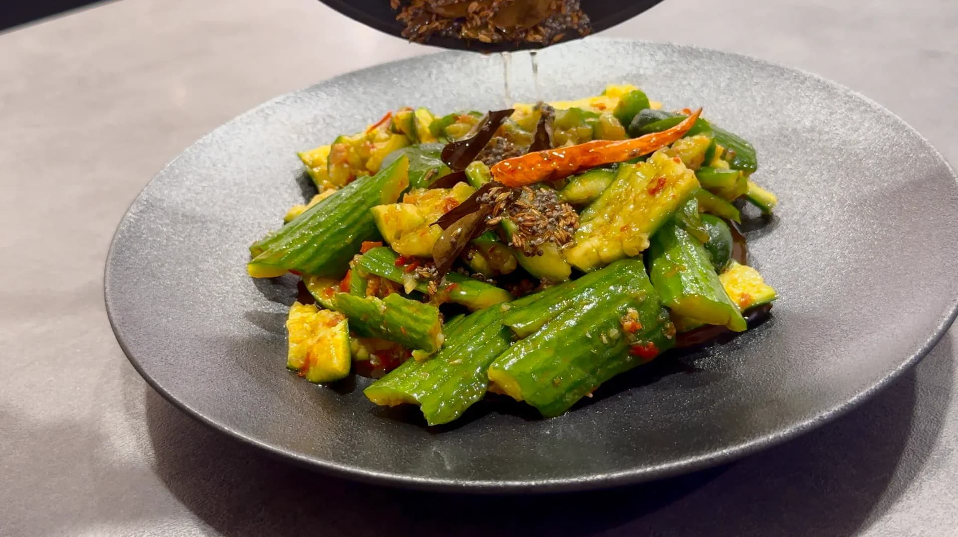 Indian fusion smashed cucumber salad with corn, green beans, and crispy tadka on a dark plate.