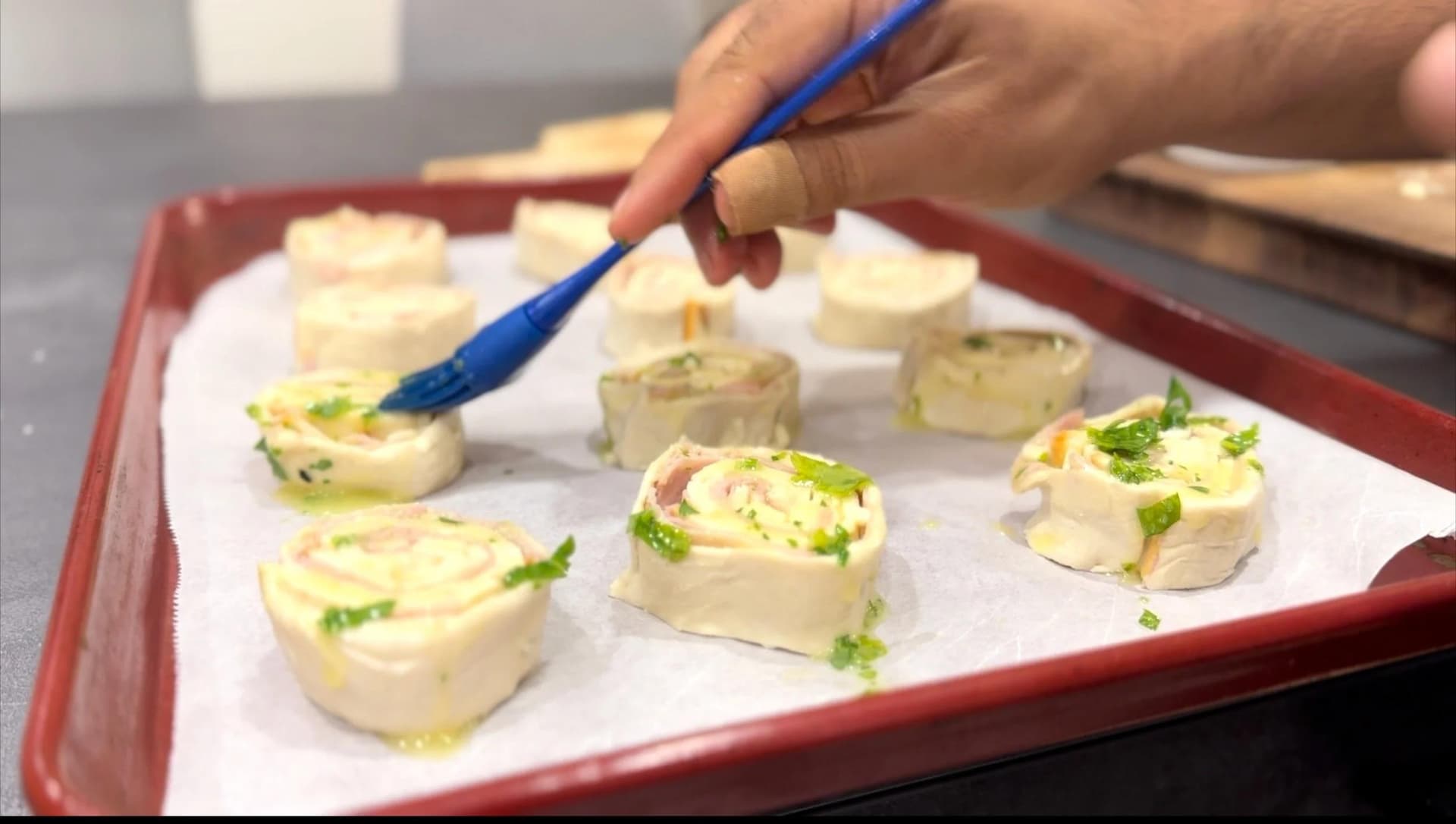 Ham and cheese puff pastry pinwheels garnished with fresh parsley on a red-bordered baking sheet.