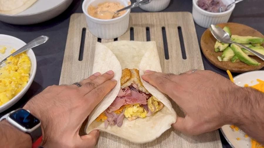Hands folding and rolling a breakfast burrito filled with scrambled eggs and ham on a wooden cutting board.