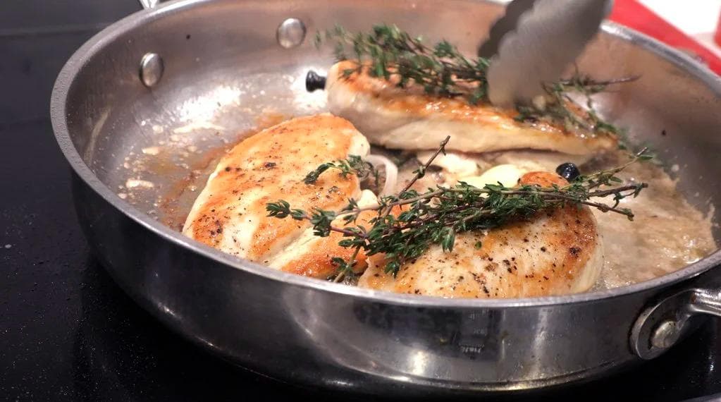 Chicken breasts being pan-fried in butter with fresh thyme sprigs and garlic cloves in a stainless steel pan.