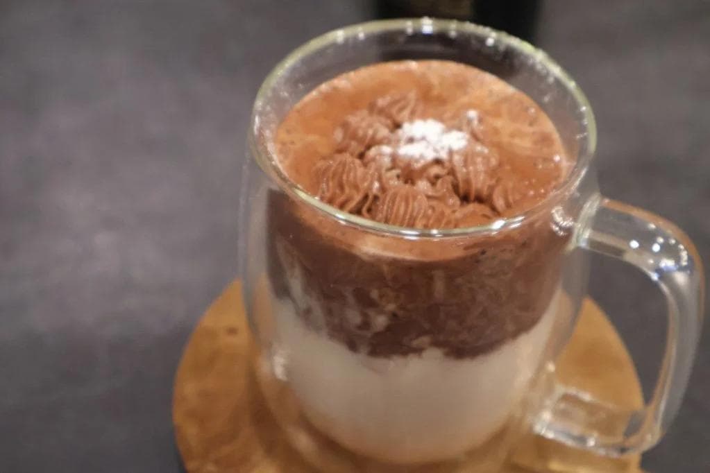 Rich whipped Baileys hot chocolate topped with cocoa powder, layered with cream in a glass mug.