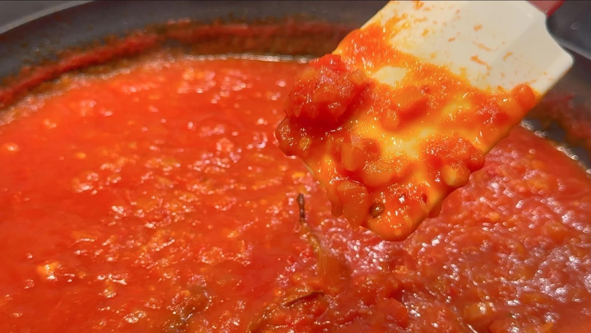 Thick, glossy red tomato piperade sauce simmering in a skillet, being stirred with a white silicone spatula.