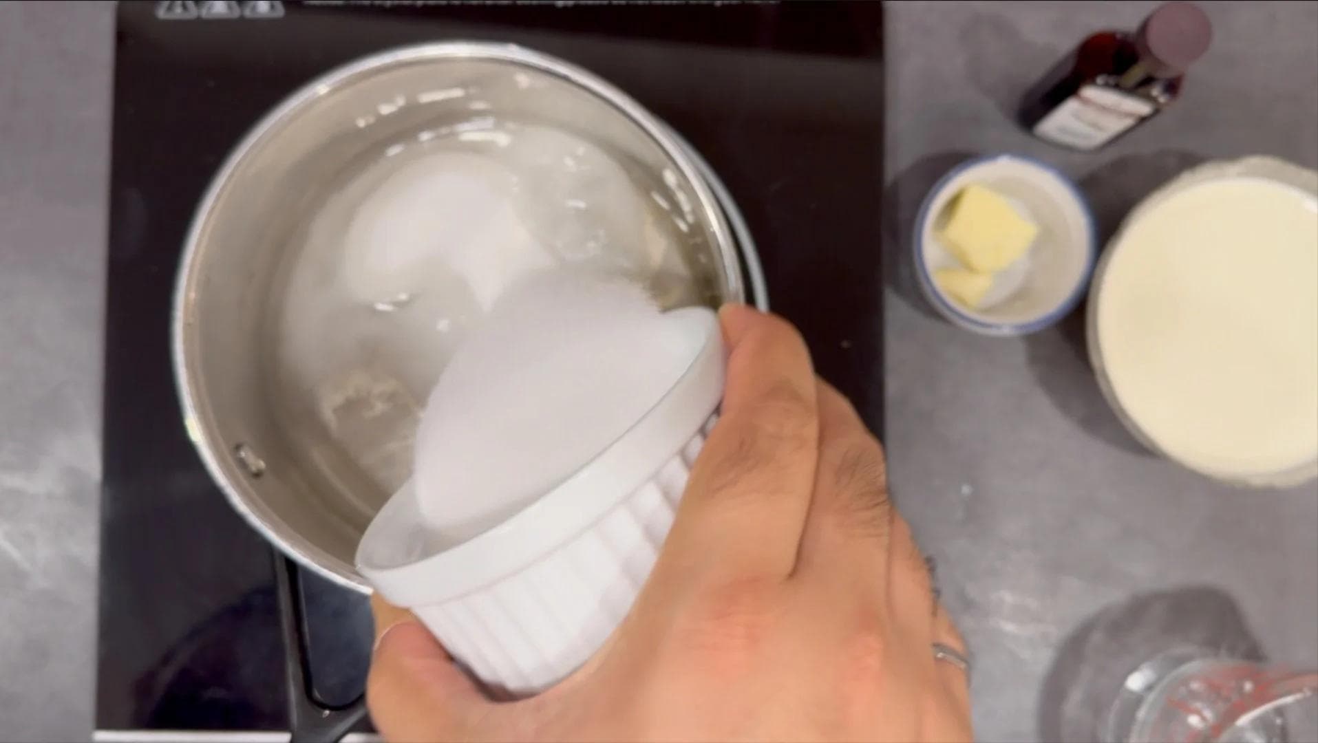 Pouring sugar and water mixture into a stainless steel pan for caramel sauce preparation.