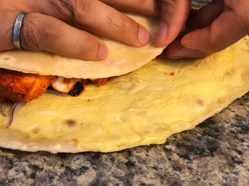 Hands wrapping a chicken tikka paratha roll with golden flatbread and spiced chicken pieces visible inside.