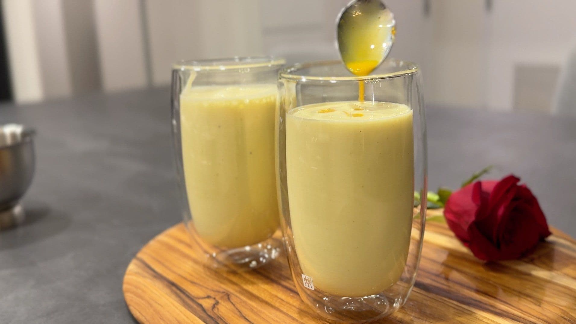 Two glasses of creamy yellow saffron and pistachio lassi with ice, garnished with crushed pistachios, on a wooden board with a red rose.
