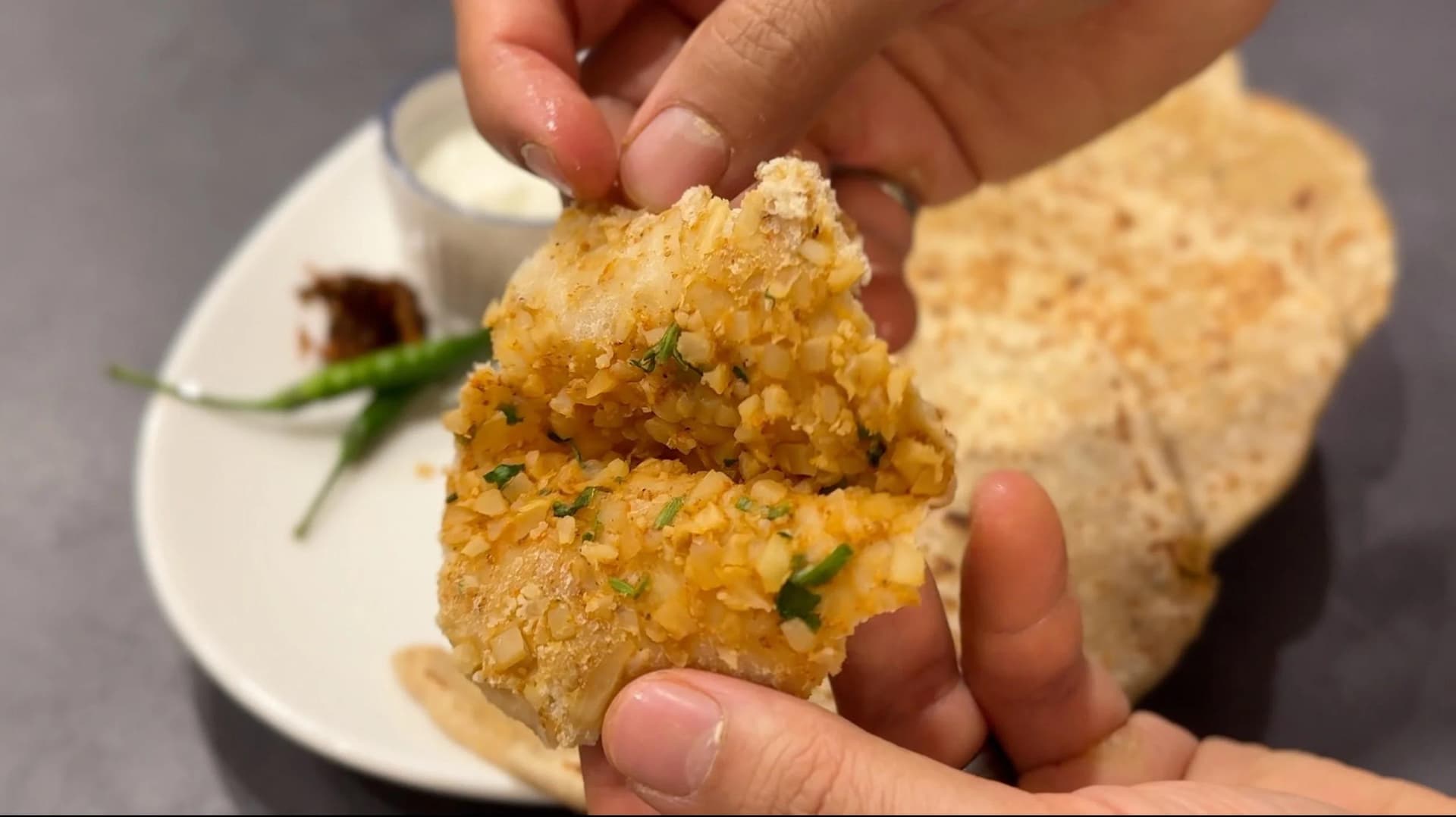 Hand holding a golden-brown cauliflower paratha garnished with fresh chives over a white plate