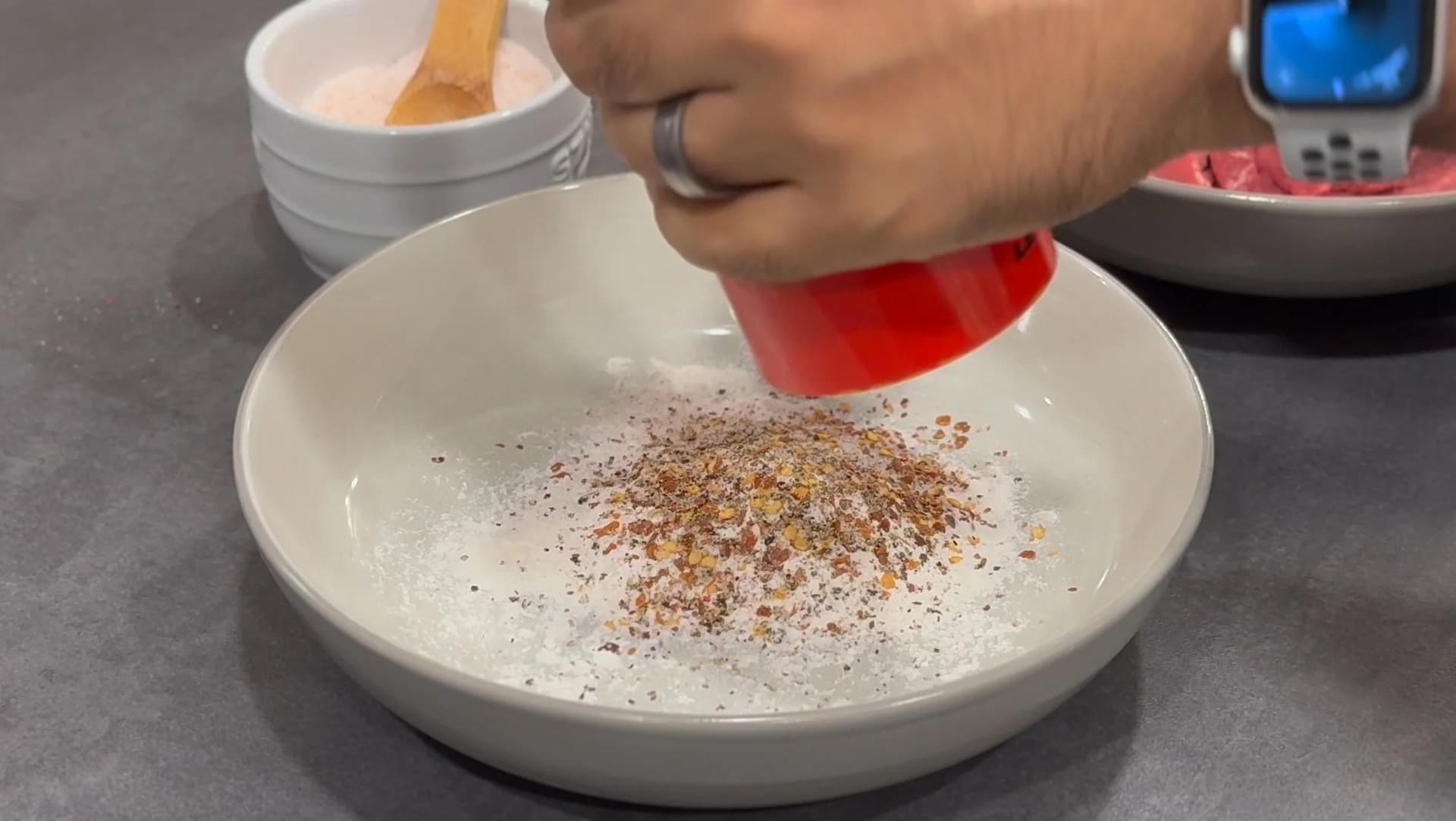 Sprinkling chili flakes and seasonings over flour in a white bowl for coating beef strips.