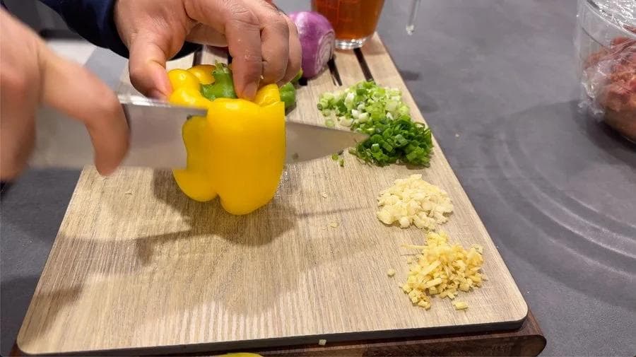 Hands slicing a yellow bell pepper on a wooden cutting board with chopped garlic, ginger, and green onions nearby.