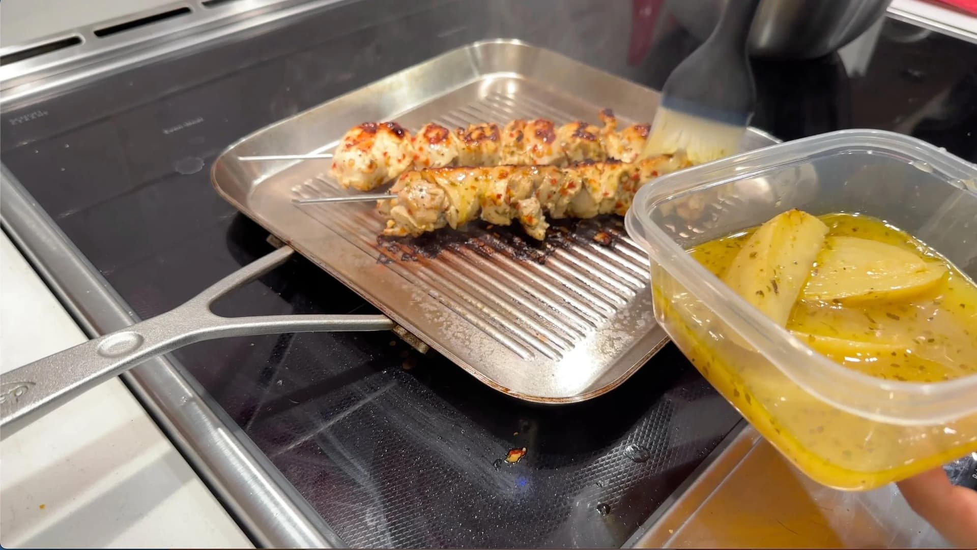 Grilled Greek souvlaki chicken skewers on a grill pan with lemon potatoes in a container beside it.