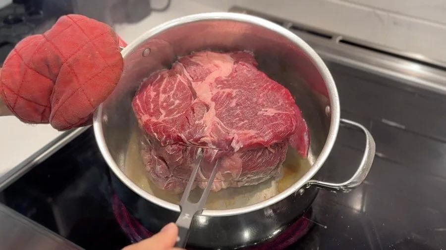 Seared beef pot roast being lifted from pot with tongs after browning, with red kitchen mitts visible nearby.
