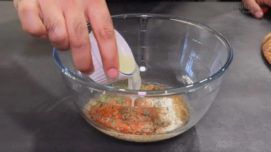 Hand pouring ghee into a glass bowl of paneer tikka marinade with spices.