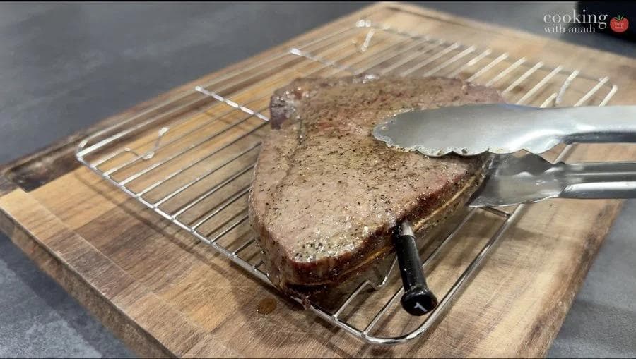 Cooked steak resting on a wire rack placed on a wooden cutting board with a meat thermometer and spatula nearby.