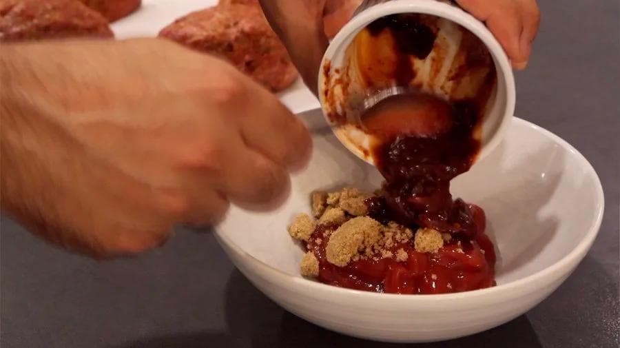 Chipotle glaze being poured from a small white bowl onto mini meatloaves topped with breadcrumb mixture in a white dish.