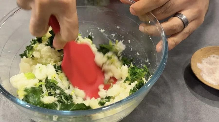 Mashing scooped potatoes with greens, butter, and cheese in a glass bowl using a red masher.