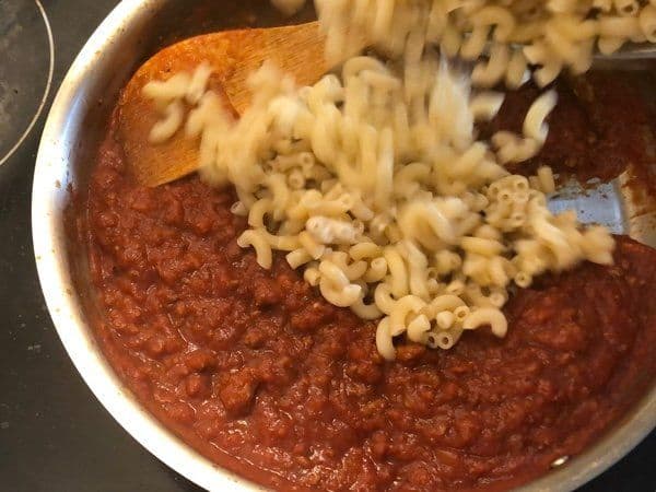 Cooked elbow pasta being added to a tomato-based meat sauce in a white pot.