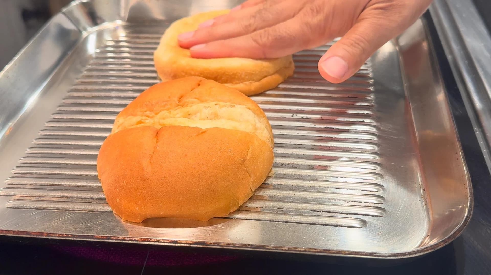 Toasted burger buns on a grill pan with a hand pressing the top bun.