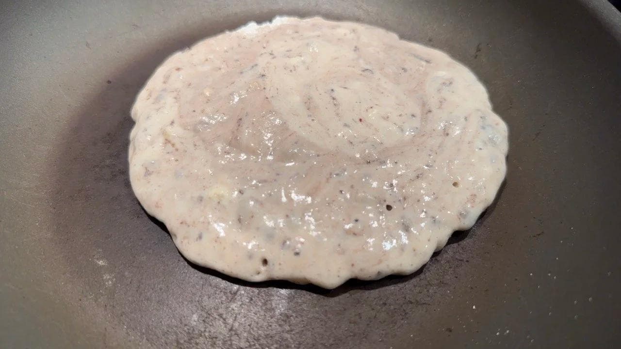 Gluten-free pancake cooking in a non-stick skillet, showing bubbles forming on the surface before flipping.