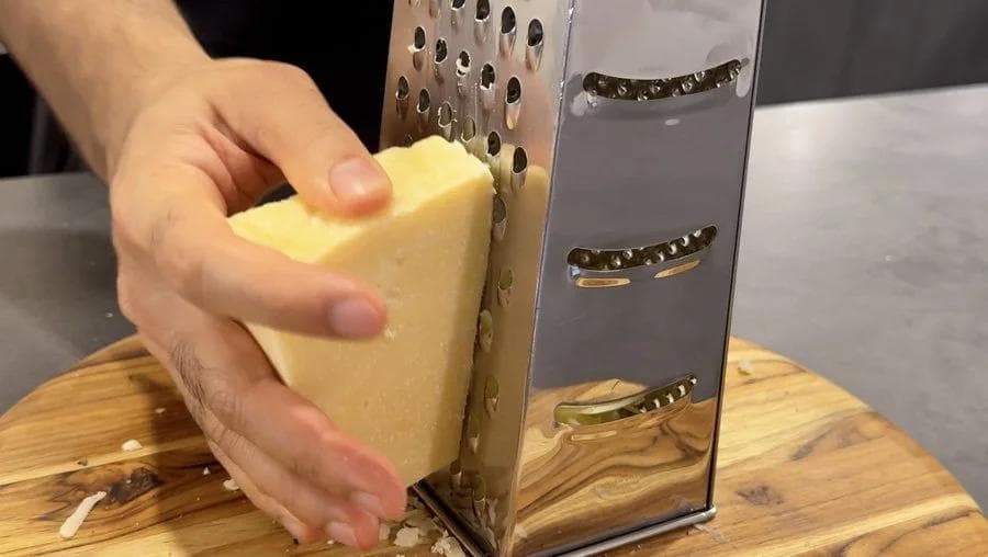 Hand grating yellow cheddar cheese over a box grater onto a wooden cutting board.