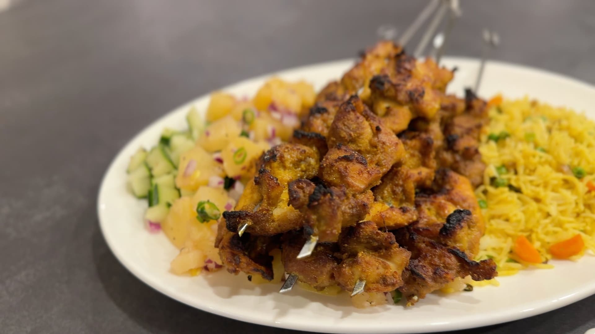 Grilled chicken satay skewers with golden-brown char served with yellow rice and diced pineapple salsa on a white plate.