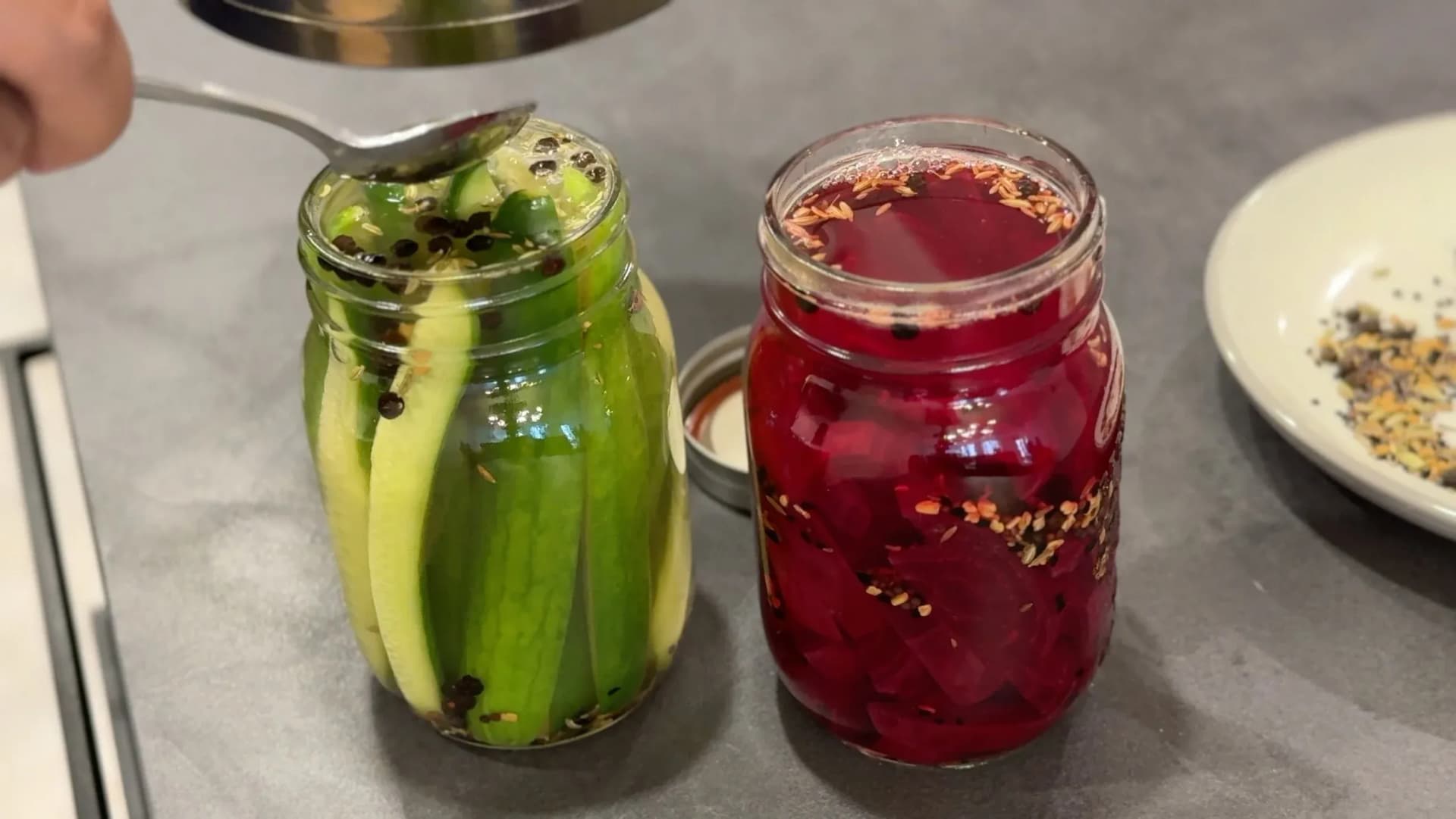 Two mason jars filled with pickling solution: one with green cucumber slices and spices, one with red pickled vegetables and spices.