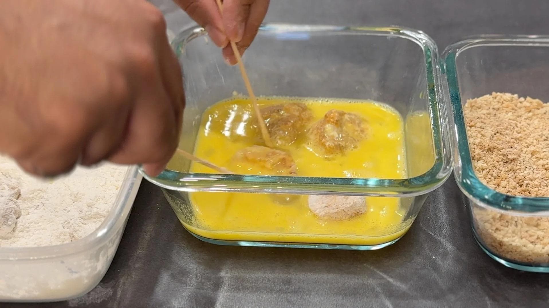 Hand dipping breaded chicken nugget into beaten egg with flour and breadcrumb bowls nearby.