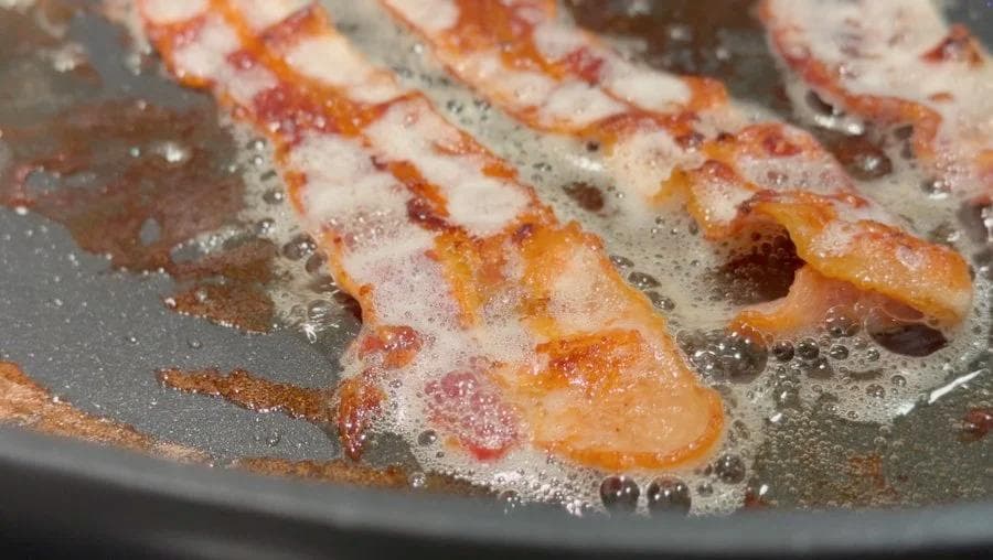 Bacon strips cooking in a pan with bubbling grease and golden-brown edges.