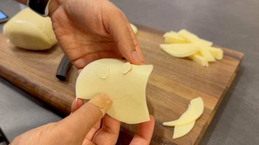 Hands cutting eye and mouth shapes from a slice of pale cheese on a wooden cutting board for ghost pizza decorations.