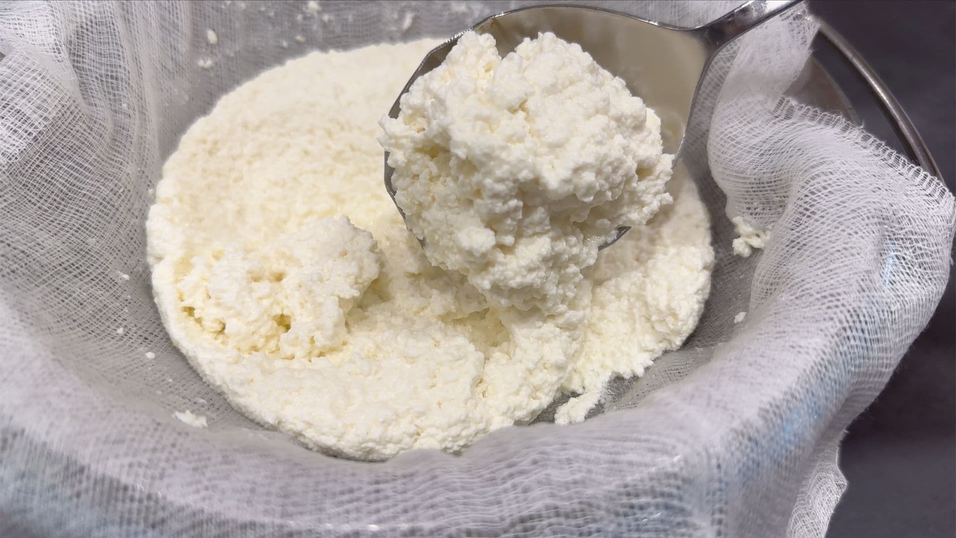 Homemade ricotta cheese curds being strained through cheesecloth with a spoon.