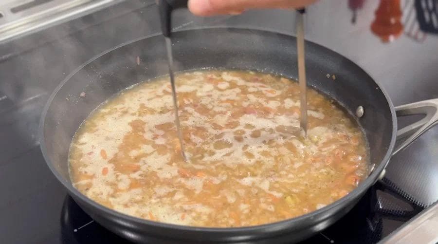 Mashing cooked beans in a simmering broth with two wooden spoons in a skillet.