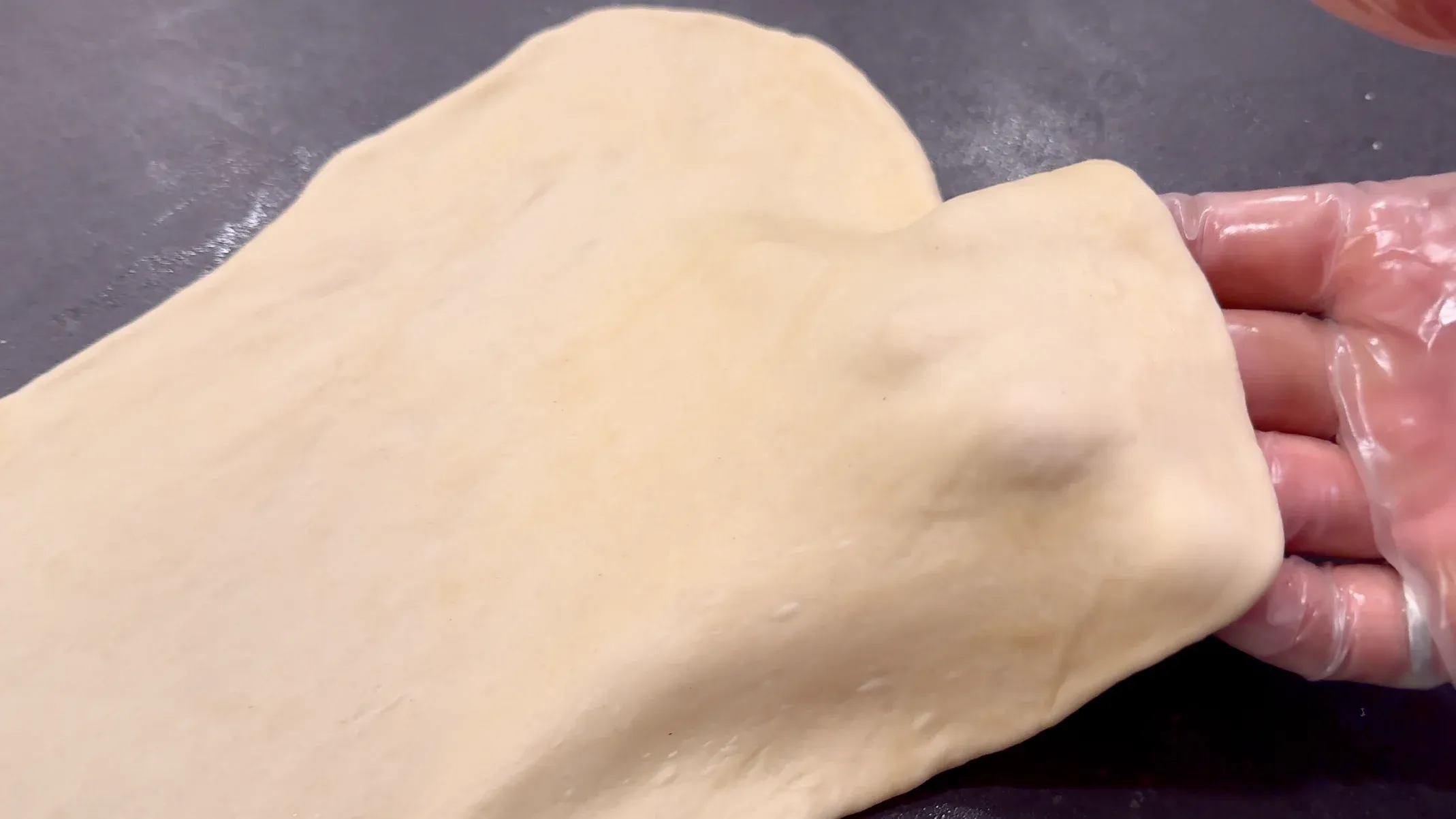 Hands stretching thin, pale Flammkuchen dough on a dark work surface.