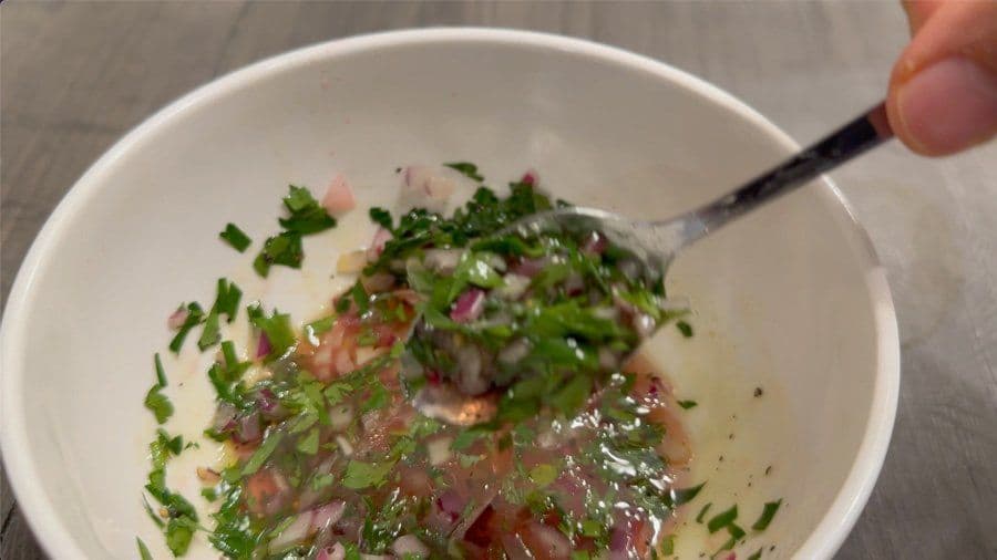 Chimichurri sauce mixed in a white bowl with chopped parsley, garlic, and red onions, with a spoon lifting a portion.