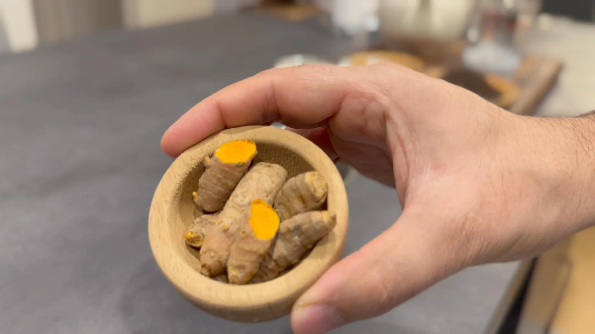 Hand holding wooden bowl with smashed turmeric root and ginger pieces topped with turmeric powder.