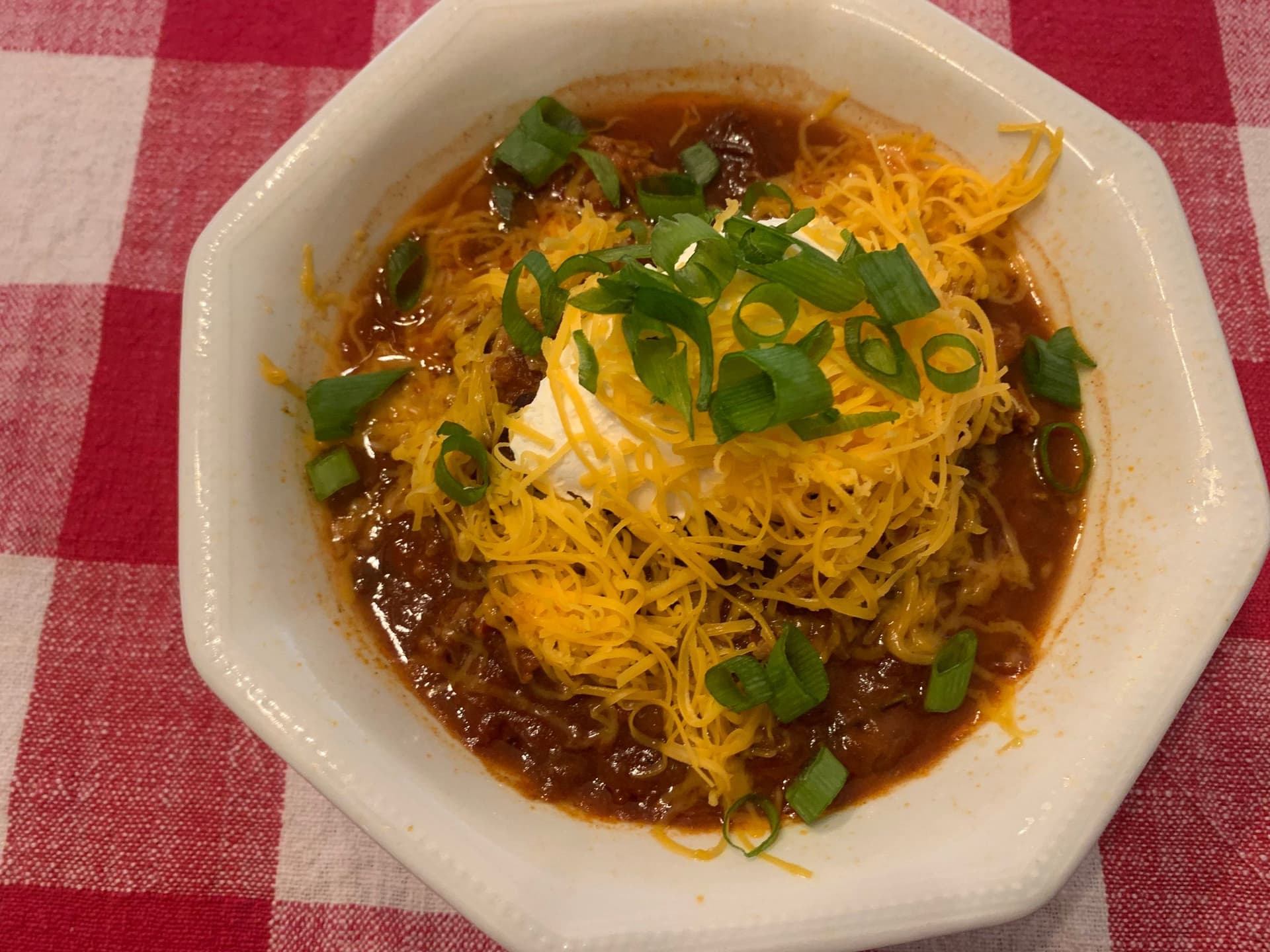 Bowl of chicken and bean chili topped with crispy chow mein noodles and sliced green onions on red gingham tablecloth.